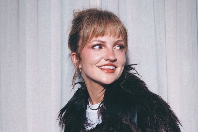 Close-up portrait of singer-songwriter Eloise with bangs and an updo hairstyle, smiling in a dark fur-trimmed jacket against a light curtain.